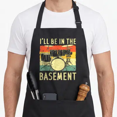 Drumming drum set apron - Retro drum kit on red yellow green stripes with I'll be in the basement text — shown on a black cotton canvas apron against a white background