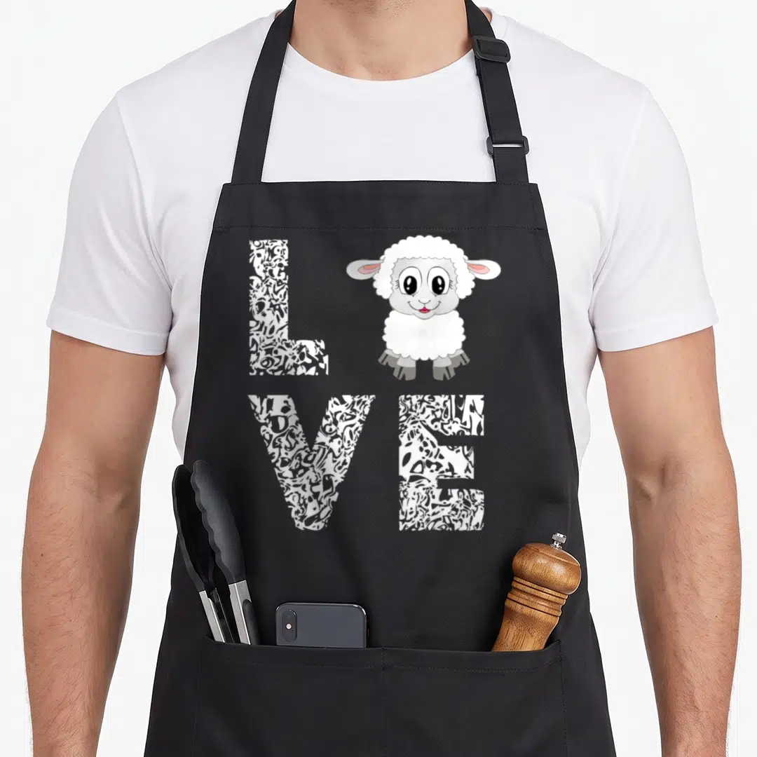Lamb sheep apron - Cartoon sheep replacing the letter O in patterned LOVE text on grey background — shown on a black cotton canvas apron against a white background