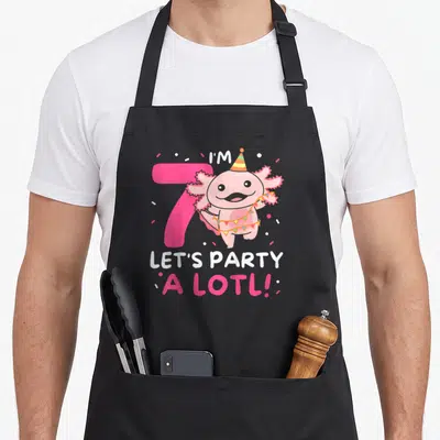 Pun birthday apron - Pink axolotl with party hat and bunting next to number 7 and text I'M 7 LET'S PARTY A LOTL! — shown on a black cotton canvas apron against a white background