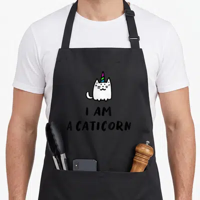 Cat unicorn apron - White cartoon caticorn with rainbow horn above stacked black text I AM A CATICORN — shown on a black cotton canvas apron against a white background