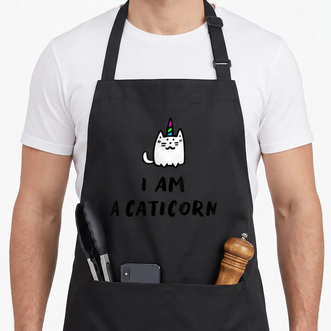 Cat unicorn apron - White cartoon caticorn with rainbow horn above stacked black text I AM A CATICORN — shown on a black cotton canvas apron against a white background