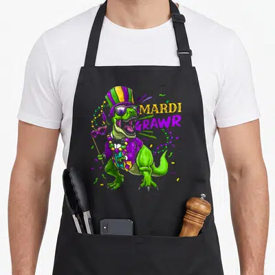 Dinosaur mardi gras aprons - Green cartoon dinosaur in purple costume holding a mask next to MARDI GRAWR text — shown on a black cotton canvas apron against a white background