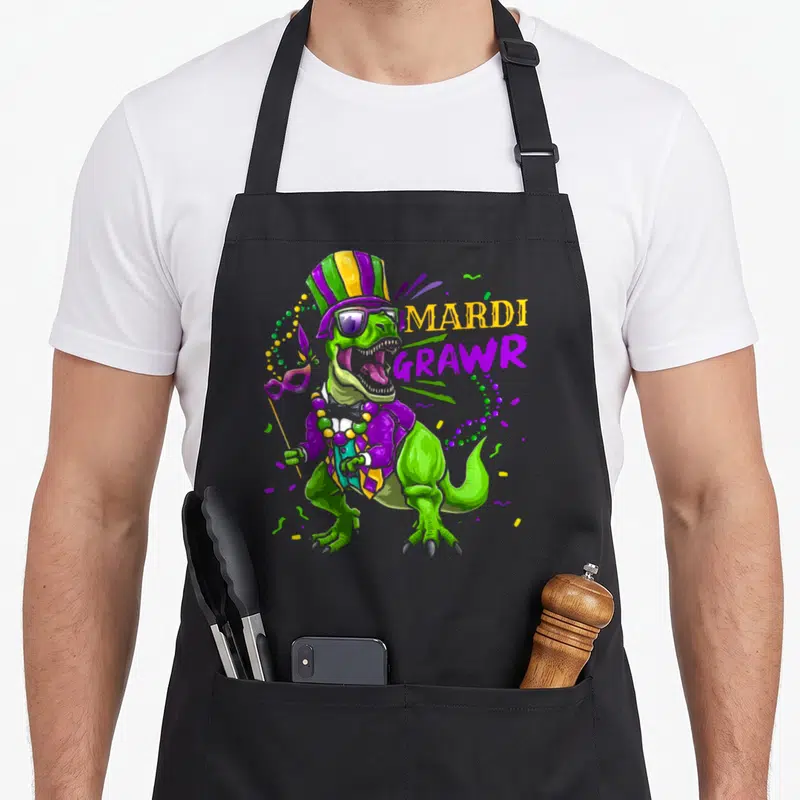 Dinosaur mardi gras aprons - Green cartoon dinosaur in purple costume holding a mask next to MARDI GRAWR text — shown on a black cotton canvas apron against a white background