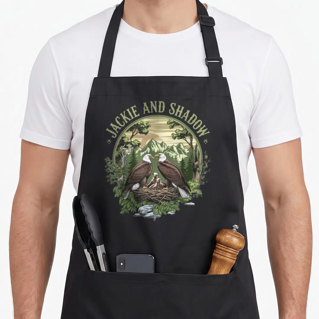 Jackie eagles apron - JACKIE AND SHADOW text over bald eagles and chick in nest with mountains — shown on a black cotton canvas apron against a white background
