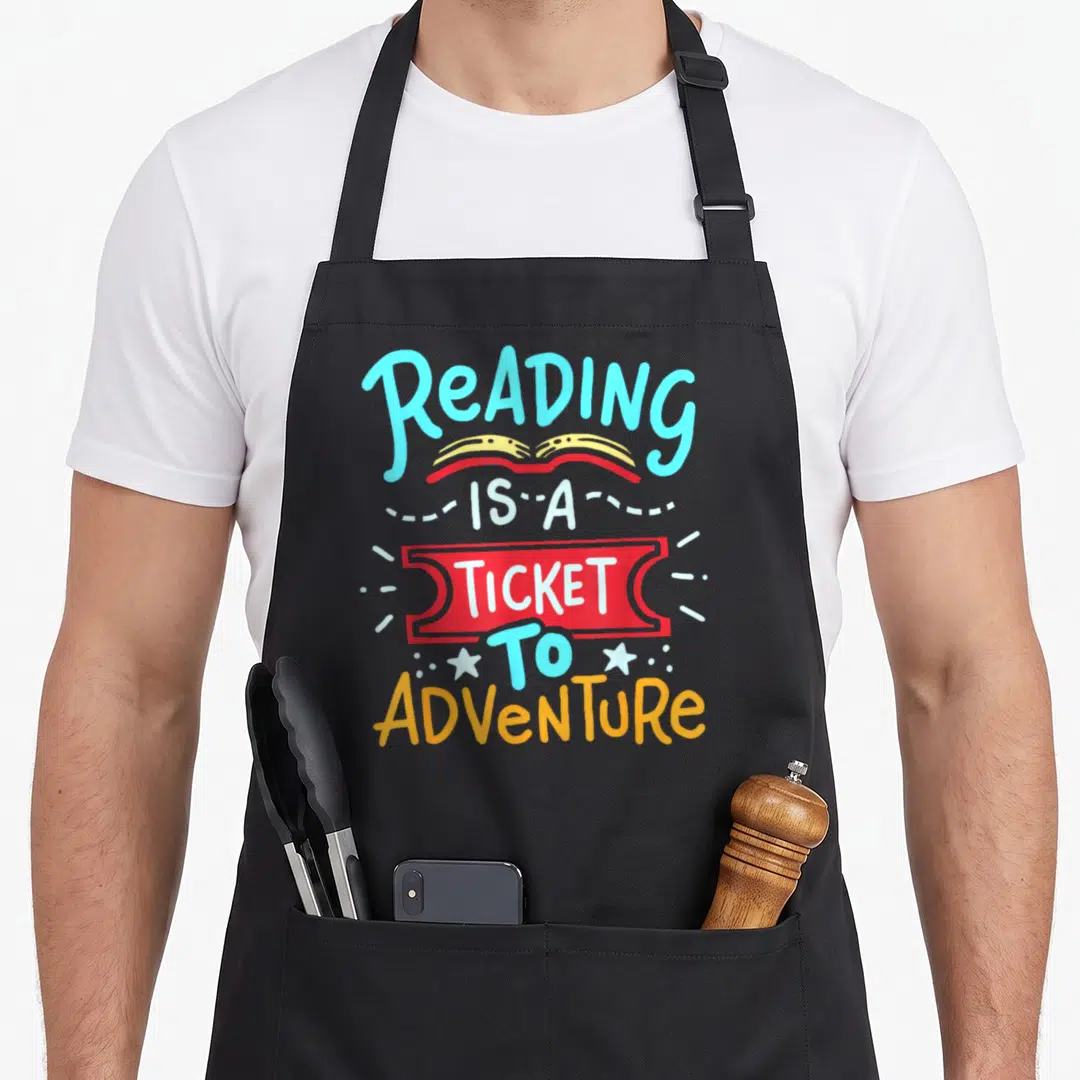 Reading teacher apron - Stacked reading quote with blue text, open book, and red ticket illustration — shown on a black cotton canvas apron against a white background