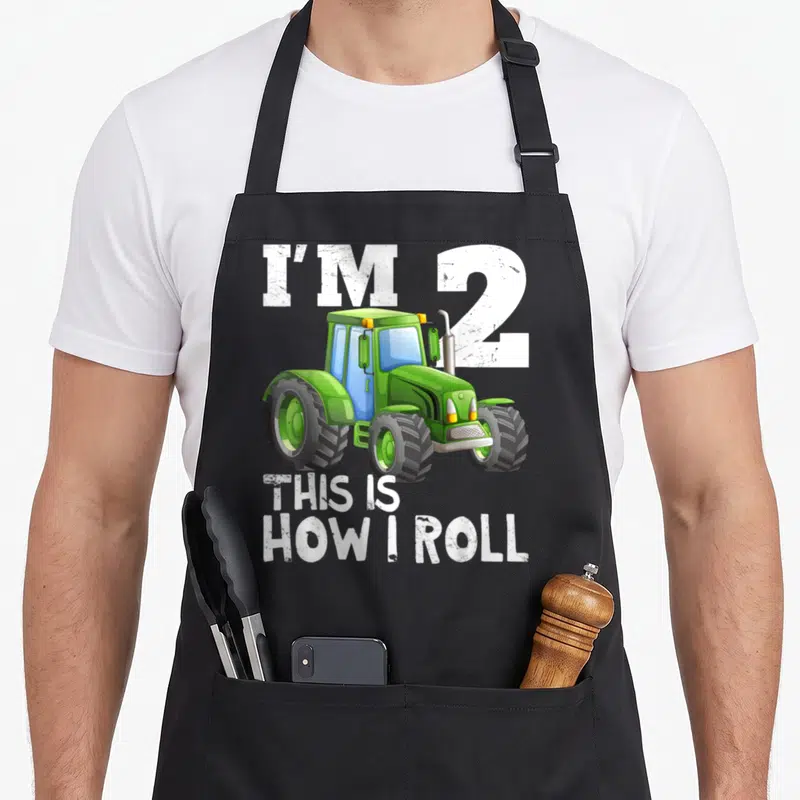 2nd farm apron - Green cartoon tractor with white text I'M 2 THIS IS HOW I ROLL — shown on a black cotton canvas apron against a white background