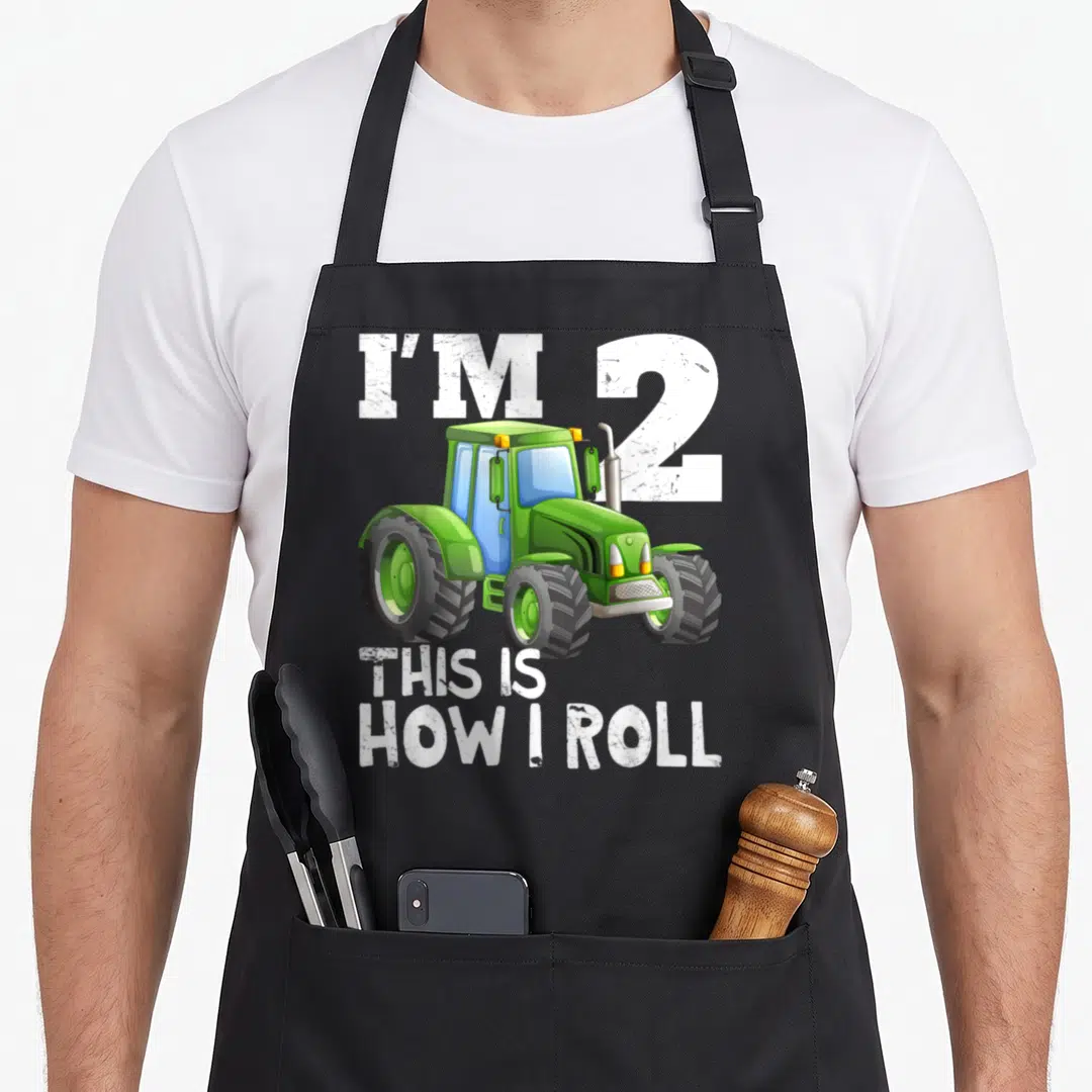 2nd farm apron - Green cartoon tractor with white text I'M 2 THIS IS HOW I ROLL — shown on a black cotton canvas apron against a white background