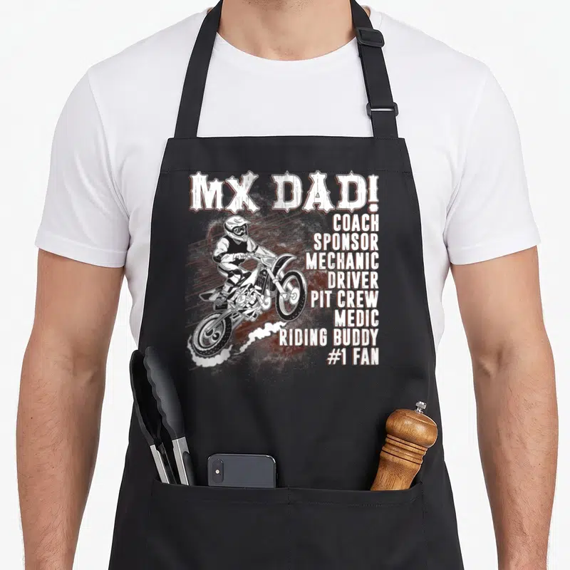 Coach bike mechanic apron - Distressed MX Dad text and motocross rider kicking up dust with stacked role titles — shown on a black cotton canvas apron against a white background