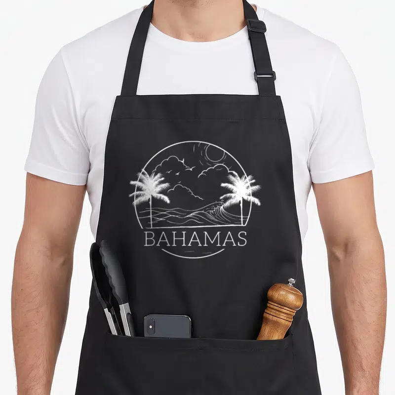 Proud bahamas flag bahamian apron - White circular line art of palm trees and ocean waves above the word BAHAMAS — shown on a black cotton canvas apron against a white background