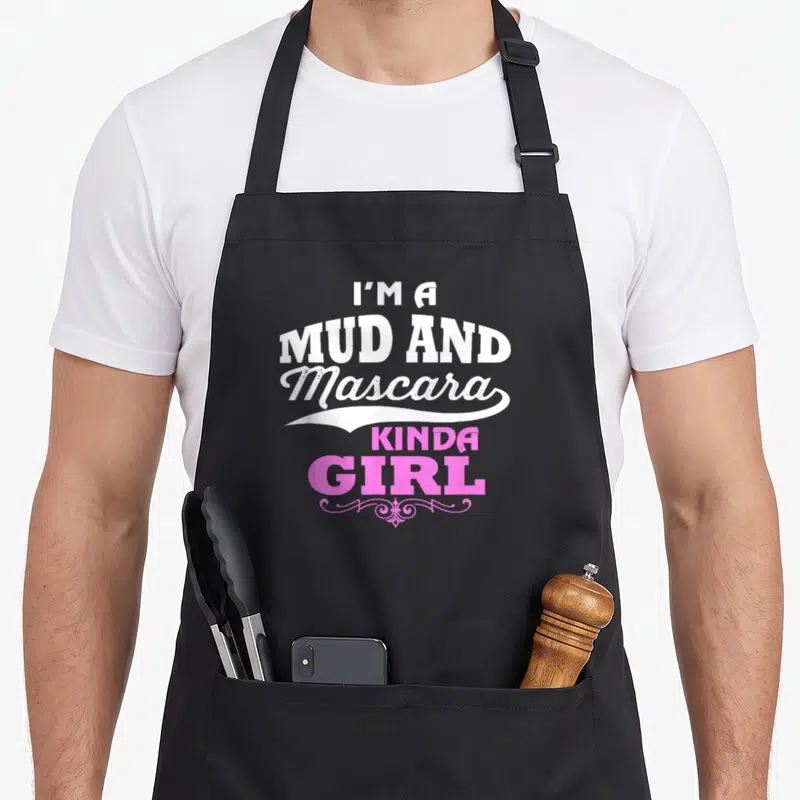 Mascara i love mud apron - Stacked white and pink text reading I'M A MUD AND Mascara KINDA GIRL with flourish — shown on a black cotton canvas apron against a white background