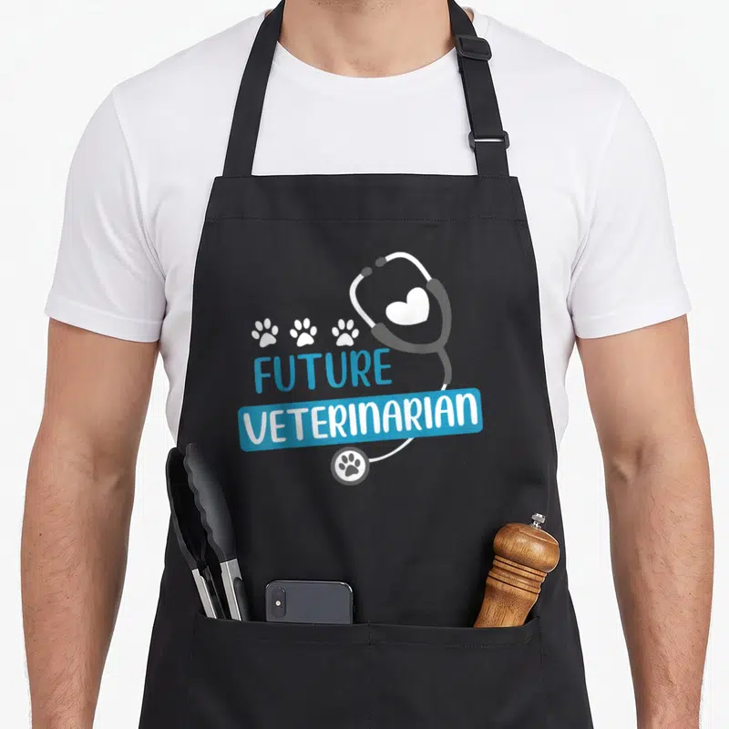 Future vet apron - Stethoscope with heart and three paw prints above Future Veterinarian text — shown on a black cotton canvas apron against a white background