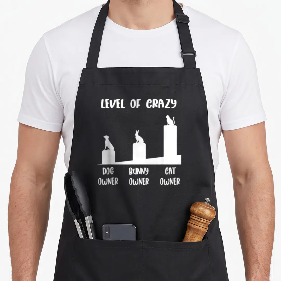 Level cat apron - White graphic of dog, bunny, and cat on rising pillars with text Level of Crazy — shown on a black cotton canvas apron against a white background