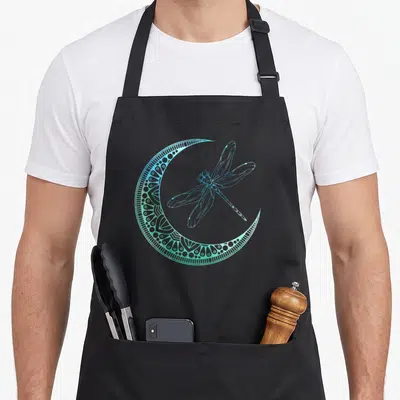Entomologist animal apron - Blue and green line-art dragonfly inside a patterned crescent moon shape — shown on a black cotton canvas apron against a white background