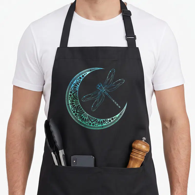 Entomologist animal apron - Blue and green line-art dragonfly inside a patterned crescent moon shape — shown on a black cotton canvas apron against a white background