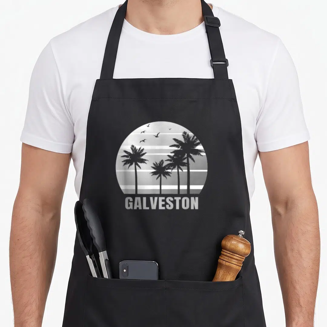 Beach texas apron - Grey and white striped circle with palm tree silhouettes and GALVESTON text below — shown on a black cotton canvas apron against a white background
