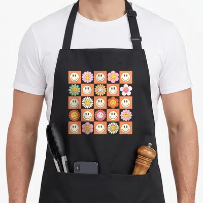 Smiling smile face on apron - Retro 5x5 grid of orange and purple squares with smiley faces and colorful daisies — shown on a black cotton canvas apron against a white background
