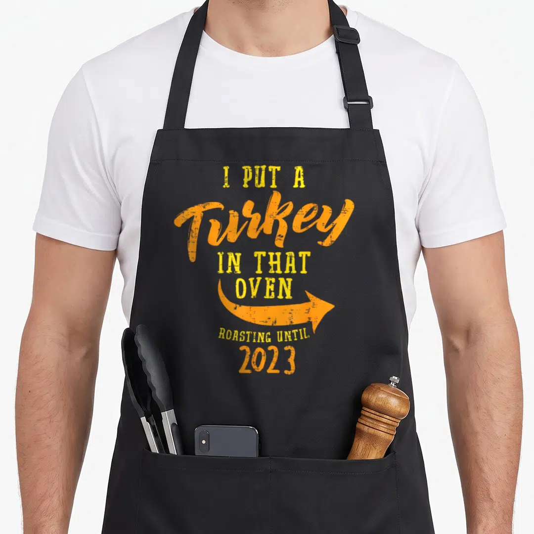 Turkey oven thanksgiving apron - Orange and yellow text reading I Put A Turkey In That Oven Roasting Until 2023 — shown on a black cotton canvas apron against a white background