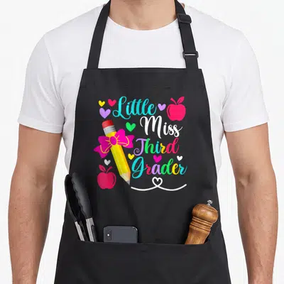 Third miss grade apron - Little Miss Third Grader text with yellow pencil, pink bow, red apples, and hearts in cyan — shown on a black cotton canvas apron against a white background