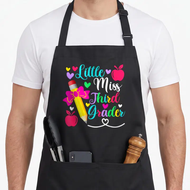 Third miss grade apron - Little Miss Third Grader text with yellow pencil, pink bow, red apples, and hearts in cyan — shown on a black cotton canvas apron against a white background
