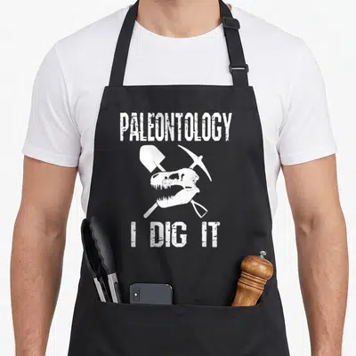 Dig science apron - White dinosaur skull with crossed shovel and pickaxe and text PALEONTOLOGY I DIG IT — shown on a black cotton canvas apron against a white background
