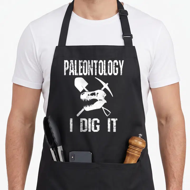 Dig science apron - White dinosaur skull with crossed shovel and pickaxe and text PALEONTOLOGY I DIG IT — shown on a black cotton canvas apron against a white background