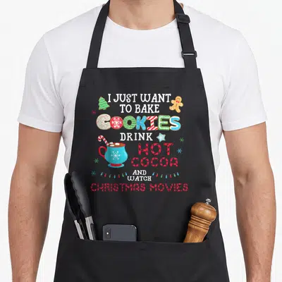 Bake watch the christmas apron - Stacked text I just want to bake cookies drink hot cocoa and watch christmas movies — shown on a black cotton canvas apron against a white background