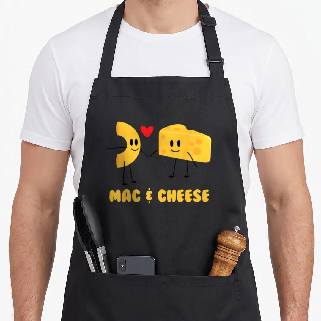 Cheese veggie apron - Cartoon macaroni noodle and cheese wedge holding hands with red heart, MAC & CHEESE text below — shown on a black cotton canvas apron against a white background