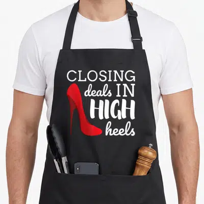 Closing realtor apron - Red high heel shoe next to white text reading closing deals in high heels — shown on a black cotton canvas apron against a white background