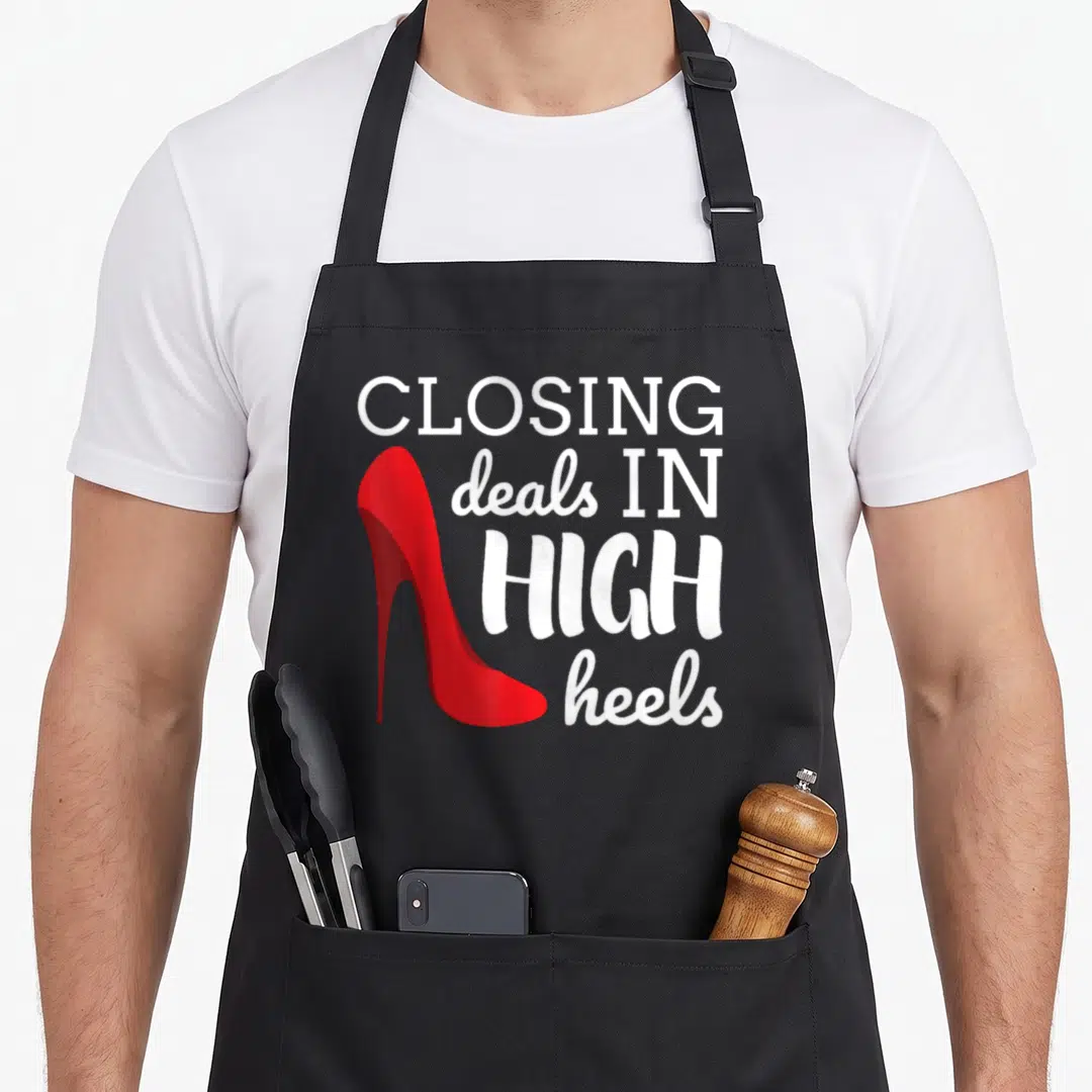 Closing realtor apron - Red high heel shoe next to white text reading closing deals in high heels — shown on a black cotton canvas apron against a white background