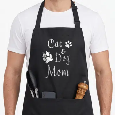 Dog kitty cat apron - White distressed text reading Cat & Dog Mom with two paw prints stacked in four lines — shown on a black cotton canvas apron against a white background