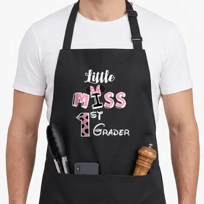 Miss hello apron - Stacked text Little MISS 1ST GRADER with mouse ears, pink polka dot bow, and dotted number 1 — shown on a black cotton canvas apron against a white background