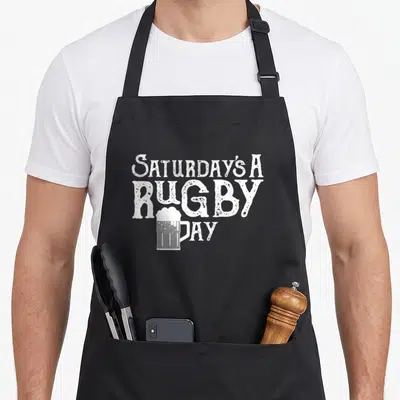 Rubgy song days apron - White distressed text Saturday's A Rugby Day with beer mug overlapping the letter D — shown on a black cotton canvas apron against a white background