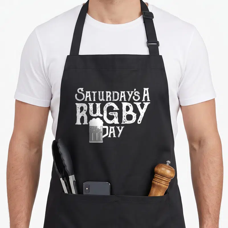 Rubgy song days apron - White distressed text Saturday's A Rugby Day with beer mug overlapping the letter D — shown on a black cotton canvas apron against a white background