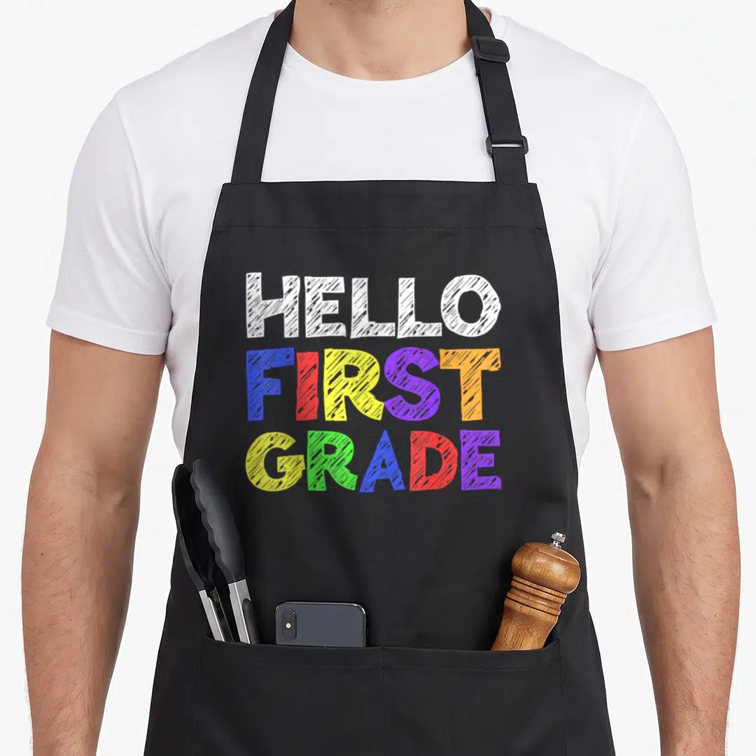 Hello teacher apron - Stacked HELLO FIRST GRADE text in white blue red yellow purple orange green sketch style — shown on a black cotton canvas apron against a white background