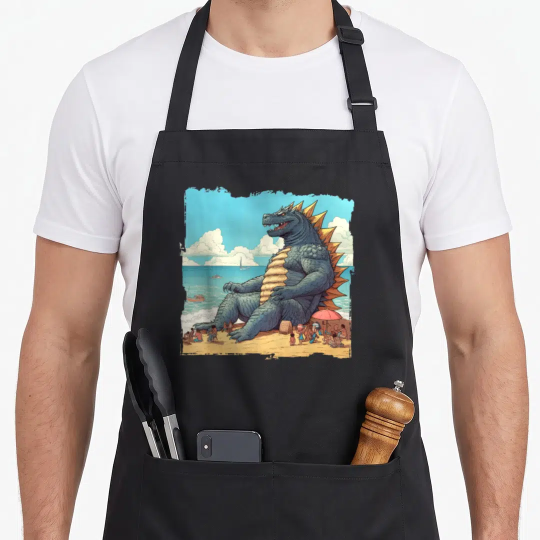 Kaiju holiday apron - Cartoon giant monster sitting on beach with tiny people and red umbrella near ocean — shown on a black cotton canvas apron against a white background