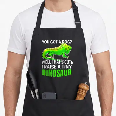 Iguana pet apron - Green cartoon iguana between white and green text reading You Got A Dog Well That Cute I Raise — shown on a black cotton canvas apron against a white background