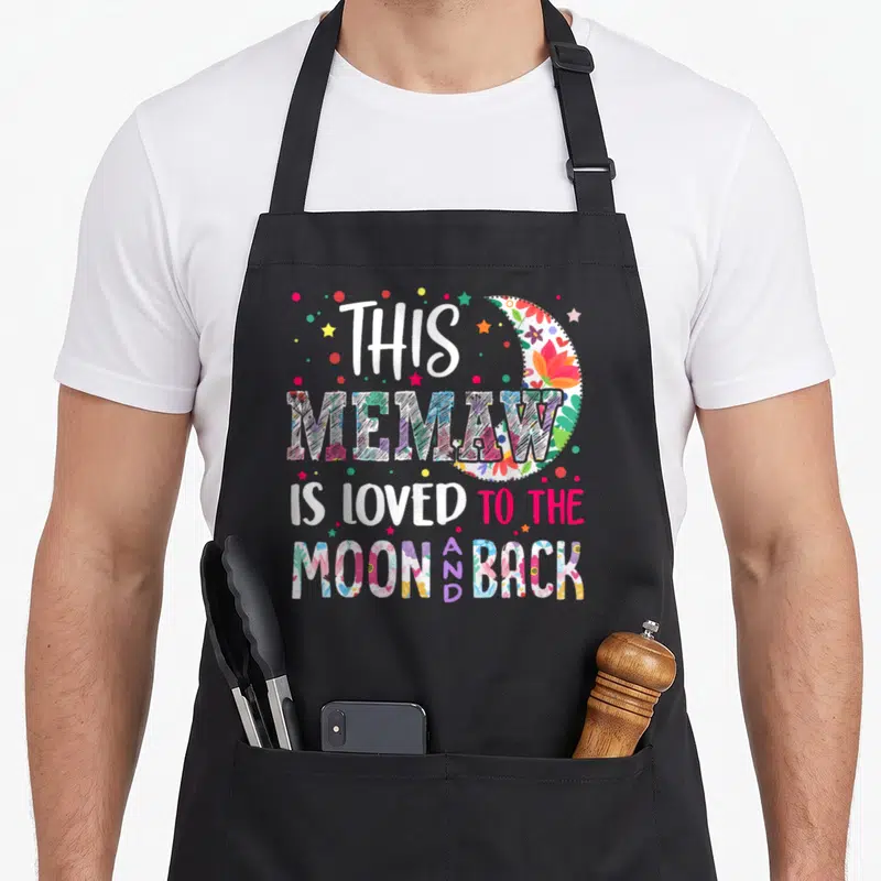 Loved memaw apron - Stacked text THIS MEMAW IS LOVED TO THE MOON AND BACK with floral moon and scattered dots — shown on a black cotton canvas apron against a white background