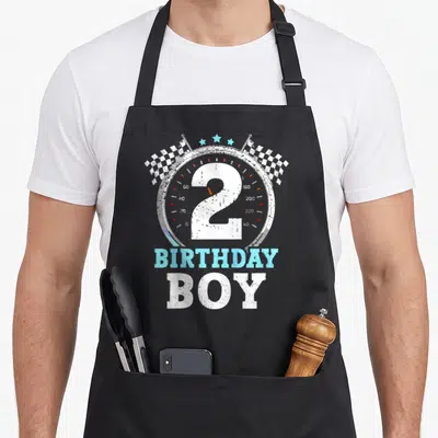 Flag 2nd apron - Speedometer with number 2, checkered flags, stars, and BIRTHDAY BOY text in distressed style — shown on a black cotton canvas apron against a white background