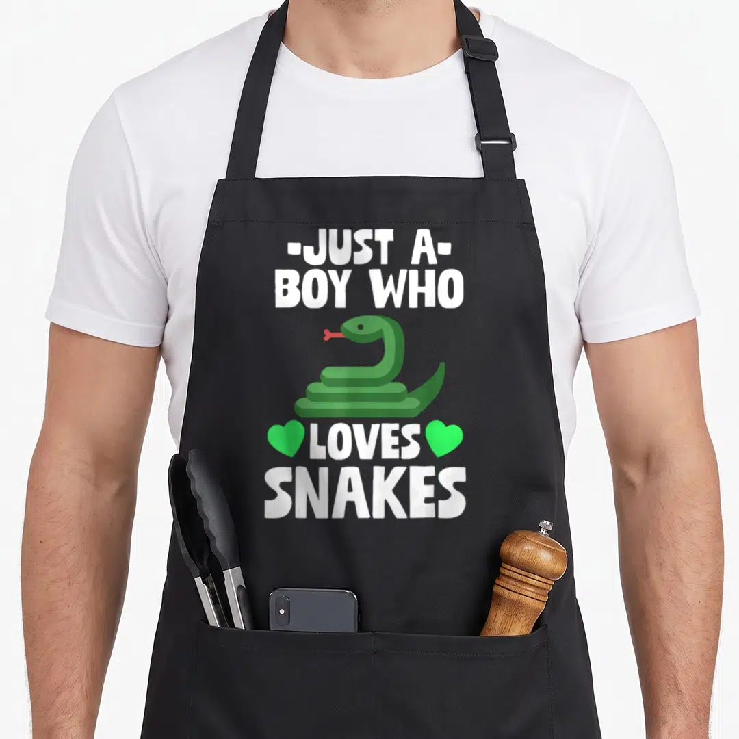 Cute snake lover apron - Green cartoon snake centered between white text Just A Boy Who Loves Snakes with two green — shown on a black cotton canvas apron against a white background