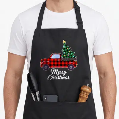 Tree buffalo apron - Red plaid truck carrying green Christmas tree with Merry Christmas script below — shown on a black cotton canvas apron against a white background
