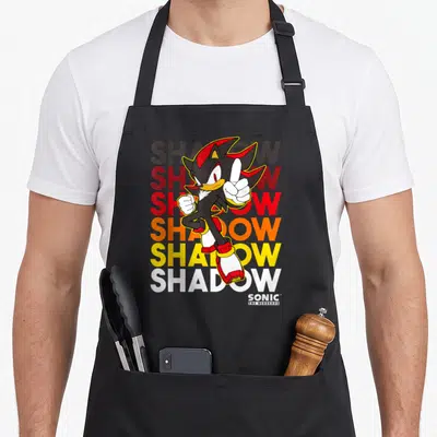 Shadow hedgehog apron - Cartoon hedgehog overlapping six stacked SHADOW words in red, orange, yellow, and white — shown on a black cotton canvas apron against a white background