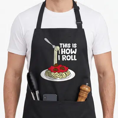 Roll pasta apron - Cartoon spaghetti plate with fork and text THIS IS HOW I ROLL — shown on a black cotton canvas apron against a white background