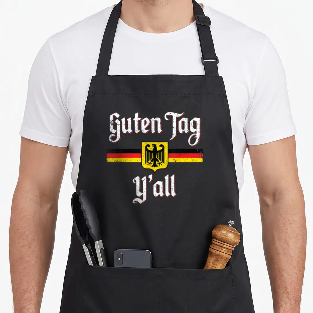 German tag apron - Guten Tag Y'all text with German eagle shield over striped flag design — shown on a black cotton canvas apron against a white background