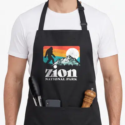 Zion bigfoot apron - Retro bigfoot silhouette over zion national park mountains against a striped red orange yellow — shown on a black cotton canvas apron against a white background