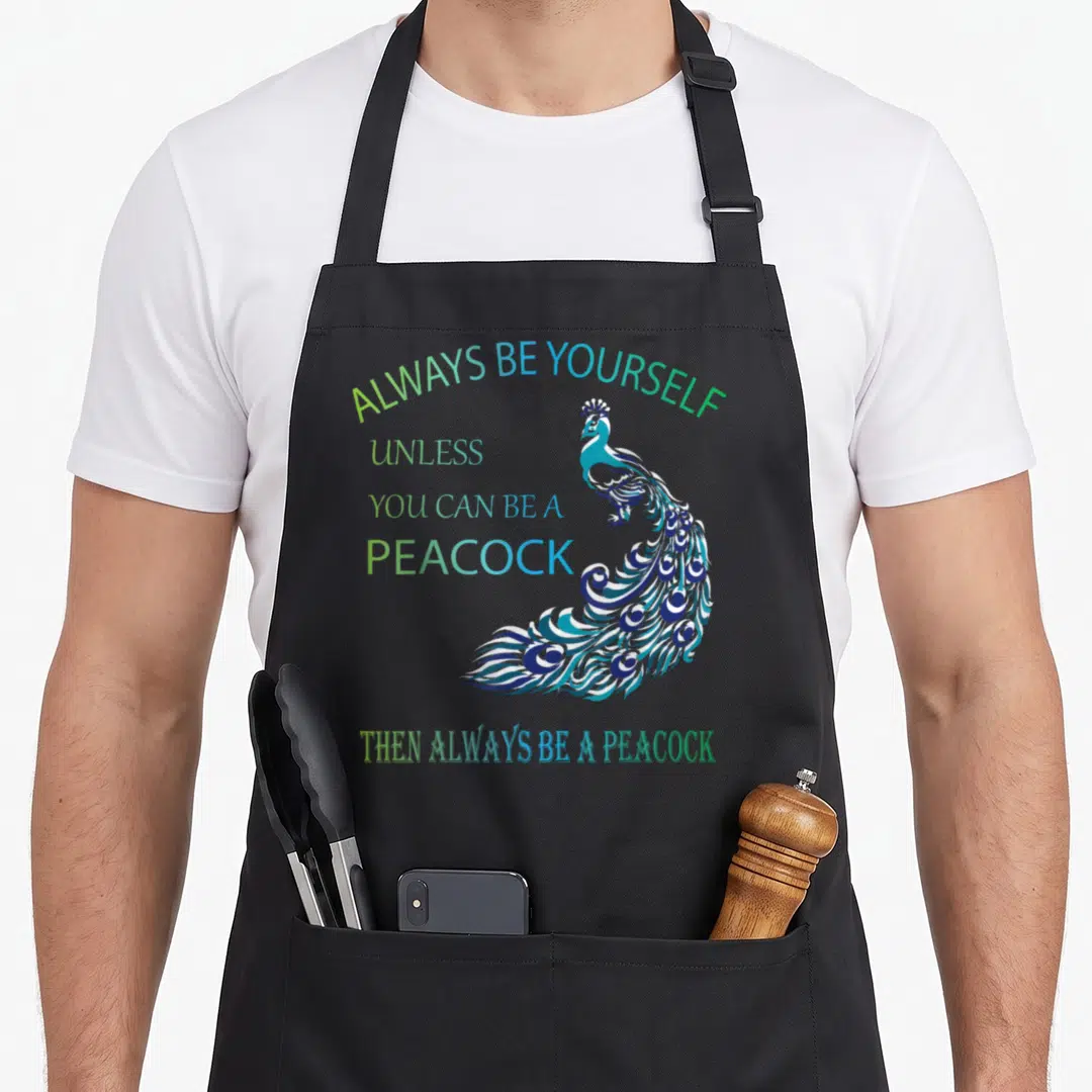 Yourself animal apron - Peacock illustration with blue green teal text Always Be Yourself unless you can be a peacock — shown on a black cotton canvas apron against a white background