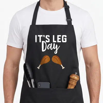 Workout thanksgiving day aprons - IT'S LEG Day text above two brown cartoon chicken drumsticks in white and brown colors — shown on a black cotton canvas apron against a white background