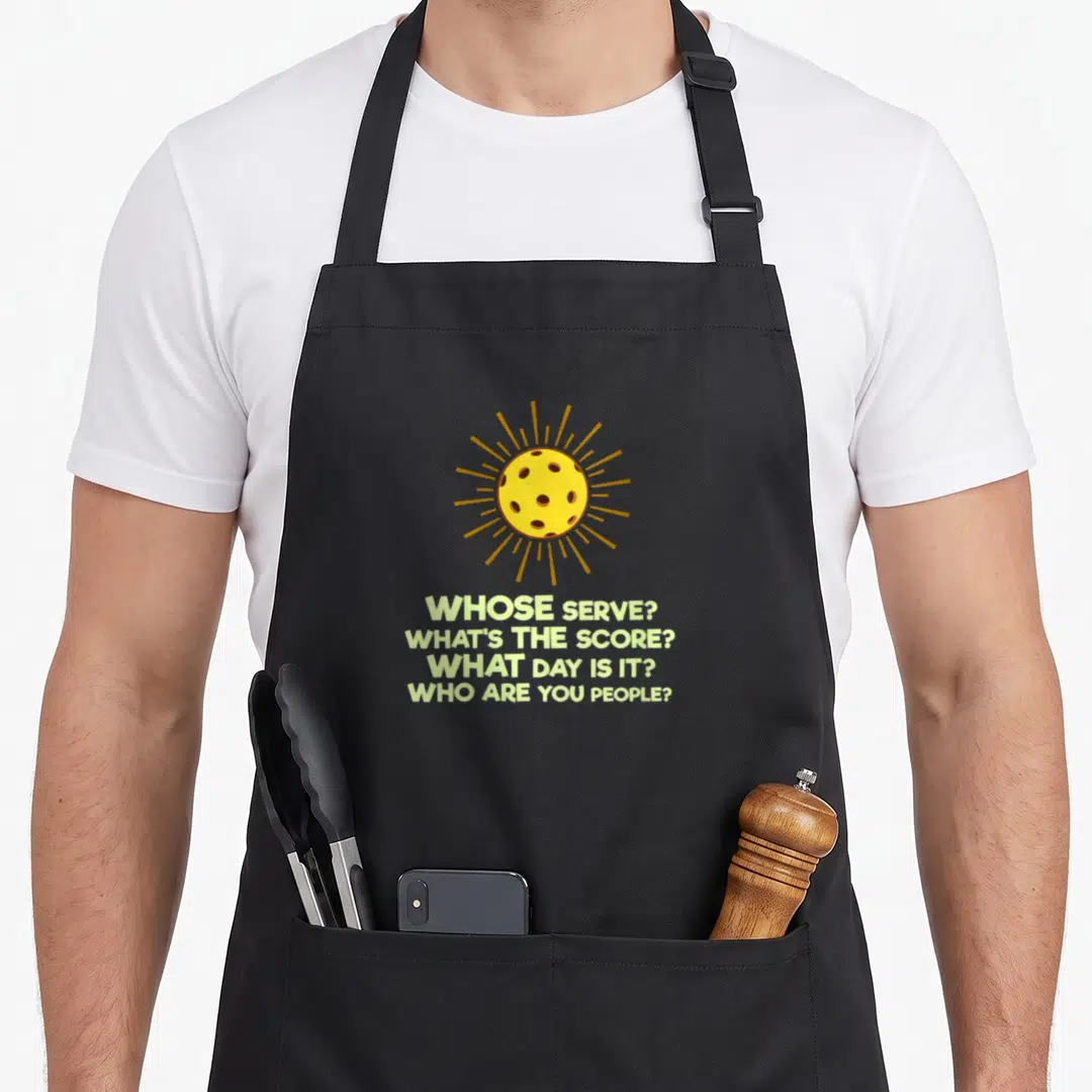 Pickleball pickle apron - Yellow ball with orange rays above four stacked questions in light green text — shown on a black cotton canvas apron against a white background