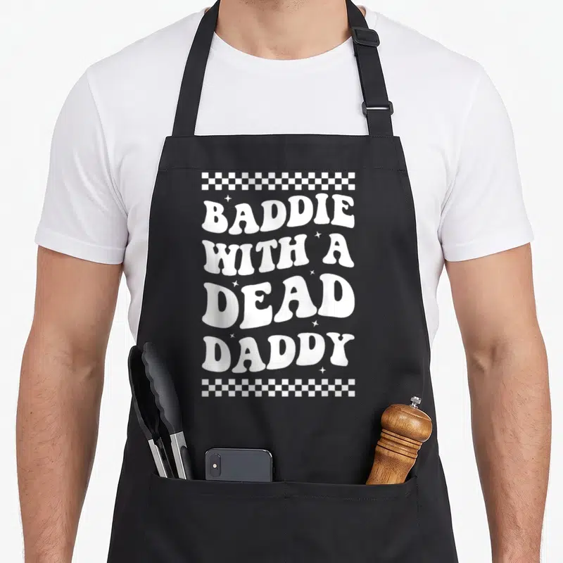 Dead daddy apron - BADDIE WITH A DEAD DADDY white text with checkerboard borders and scattered stars — shown on a black cotton canvas apron against a white background