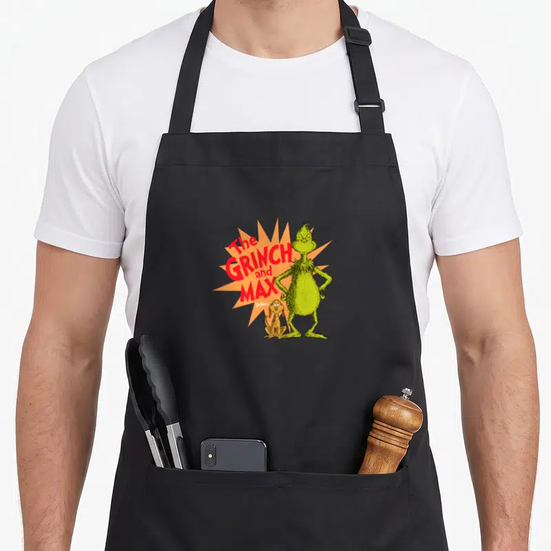 Retro max holiday apron - The GRINCH and MAX text with green character and dog on orange starburst — shown on a black cotton canvas apron against a white background