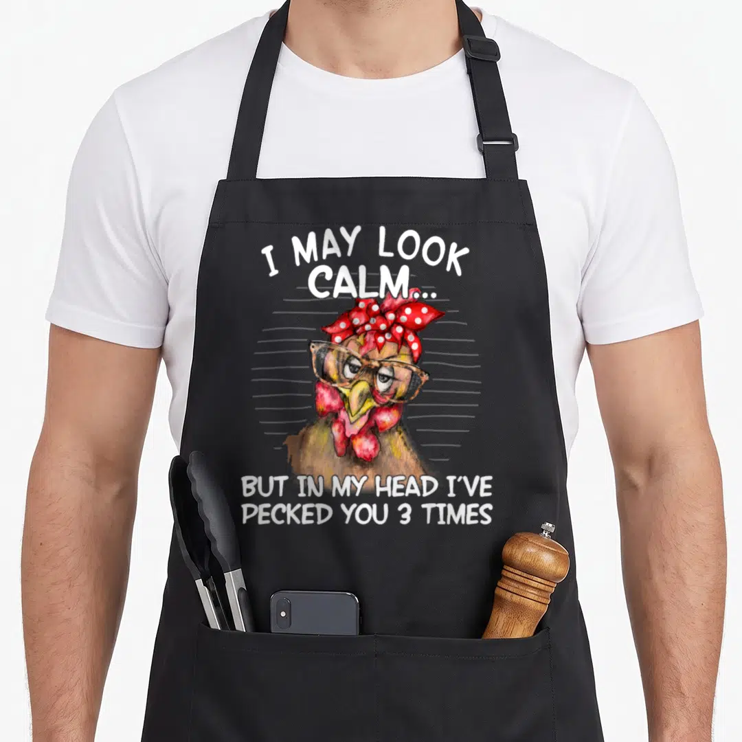 May i love my chicken apron - Cartoon chicken with glasses and red bandana, white text reading I may look calm — shown on a black cotton canvas apron against a white background